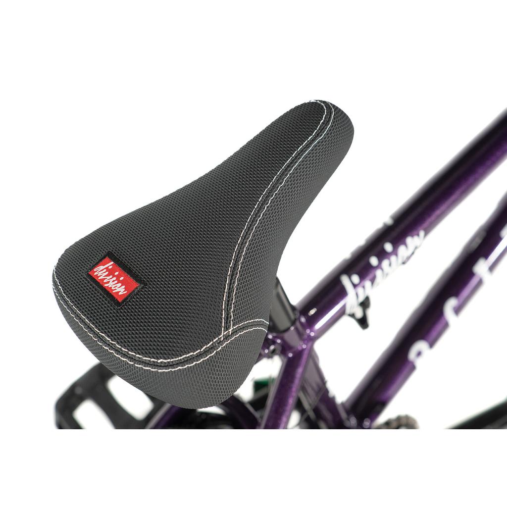 Close-up of a black textured seat with white stitching, featuring a red and white logo tag, attached to the Division Blitzer 18 Inch Bike—a purple BMX with a Hi-Ten frame.