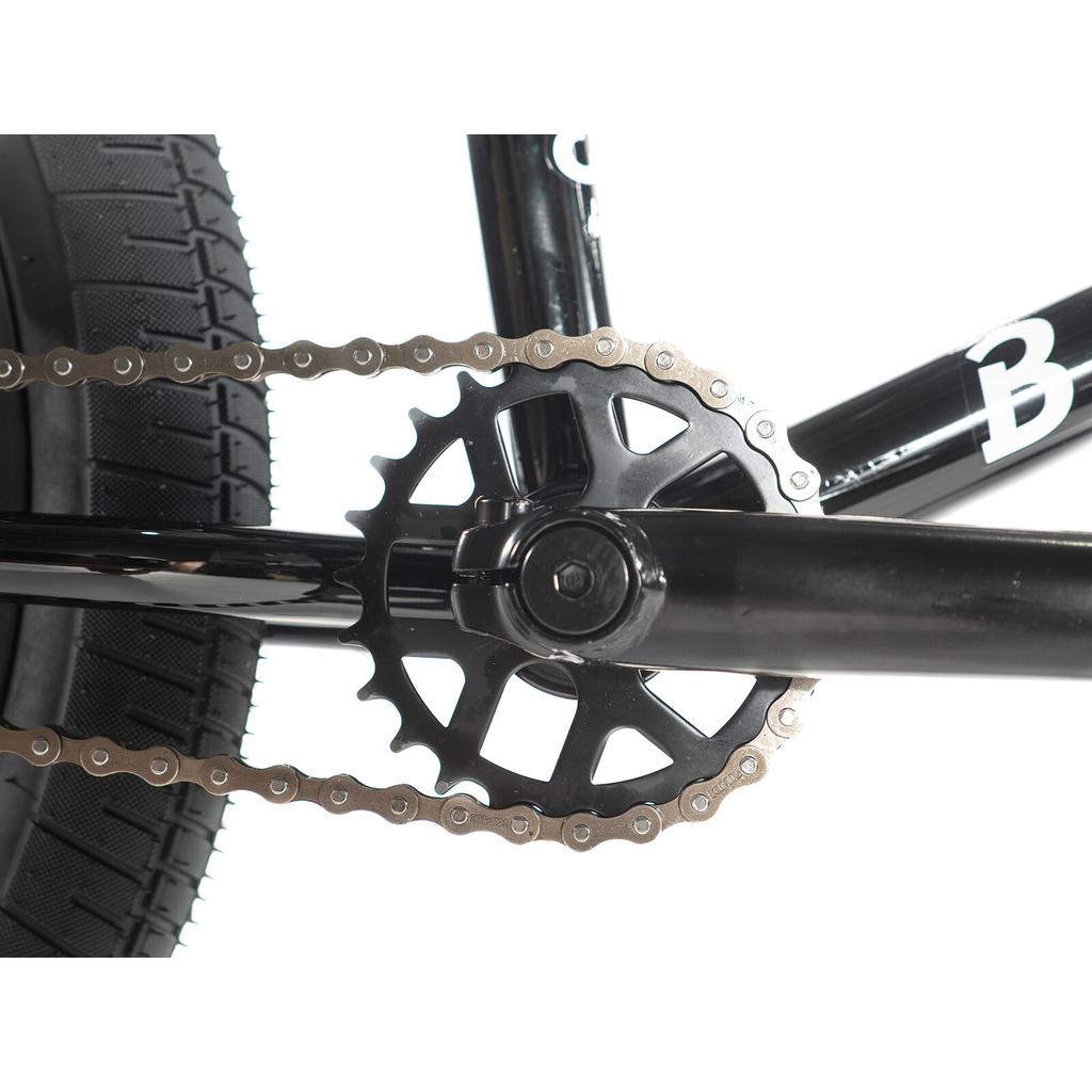 Close-up of the Division Blitzer 20 Inch Bike’s black BMX crankset, chainring, and chain, with parts of the 20” wheels, rear tire, and frame visible.