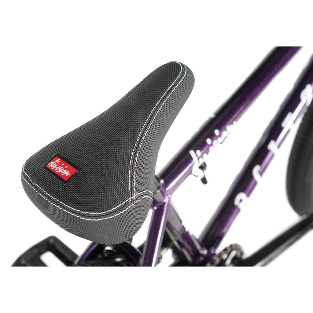 Close-up of a black textured BMX seat with white stitching and a red tag, mounted on the purple frame of the Division Blitzer 20 Inch Bike with 20" wheels.