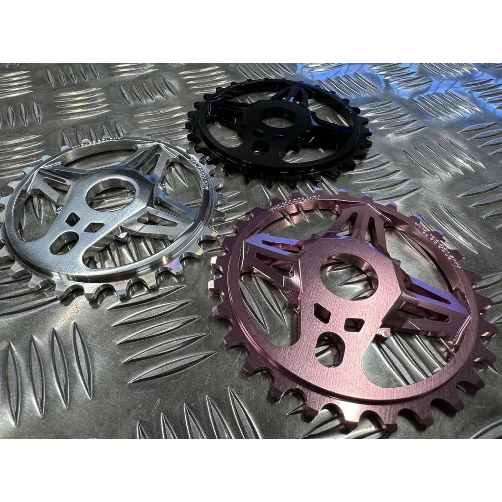 Three Colony CC Sprocket chainrings—silver, black, and purple—made from 7075T6 Alloy are displayed on a diamond plate-textured metal surface.