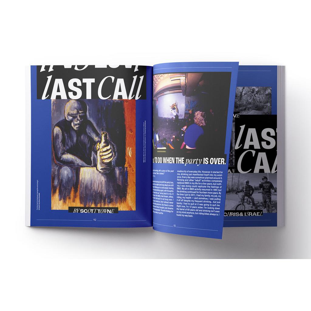 An open Greystoke Magazine V2 rests on a blue background, showing a painting of a person holding a bottle on the left page and text with two small photos on the right.