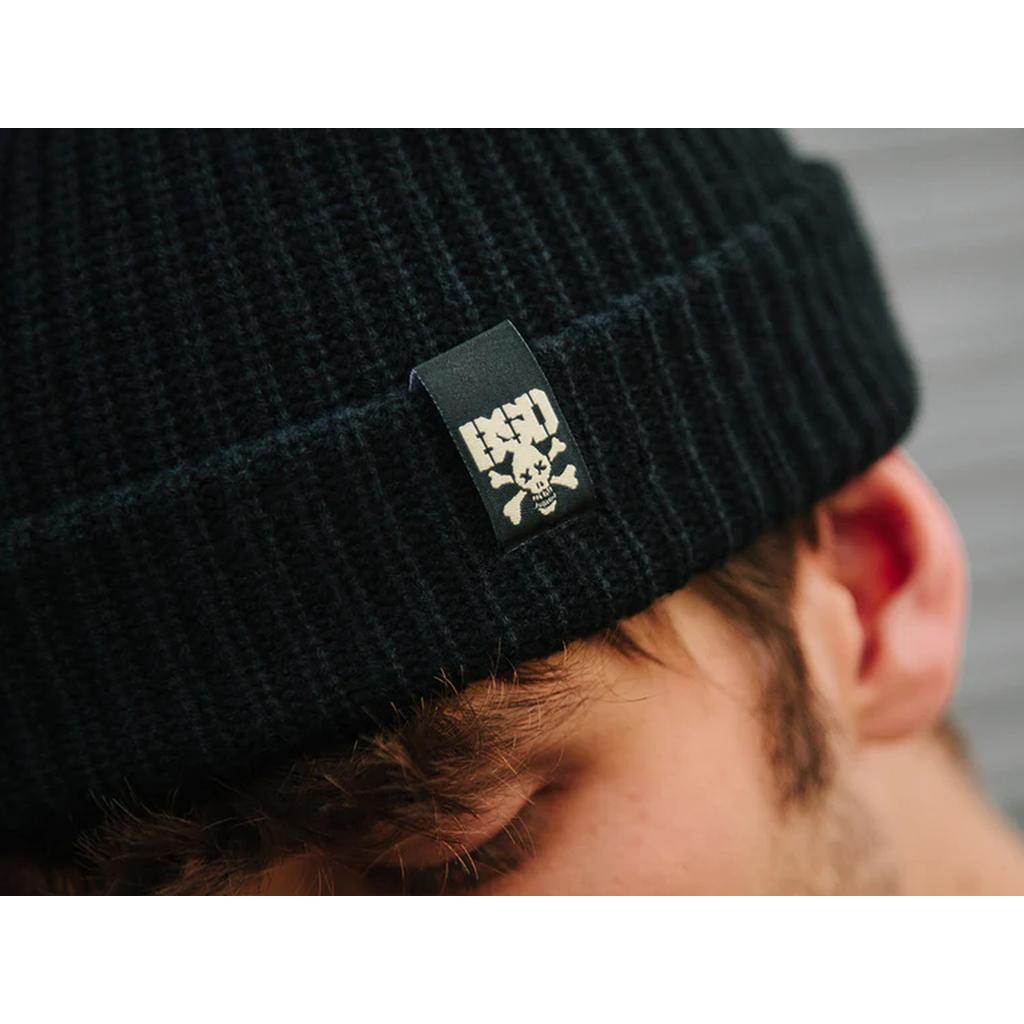 A close-up of a person wearing the BSD More Speed Beanie, which features a wide knit design and a tag with a white skull and crossbones graphic.