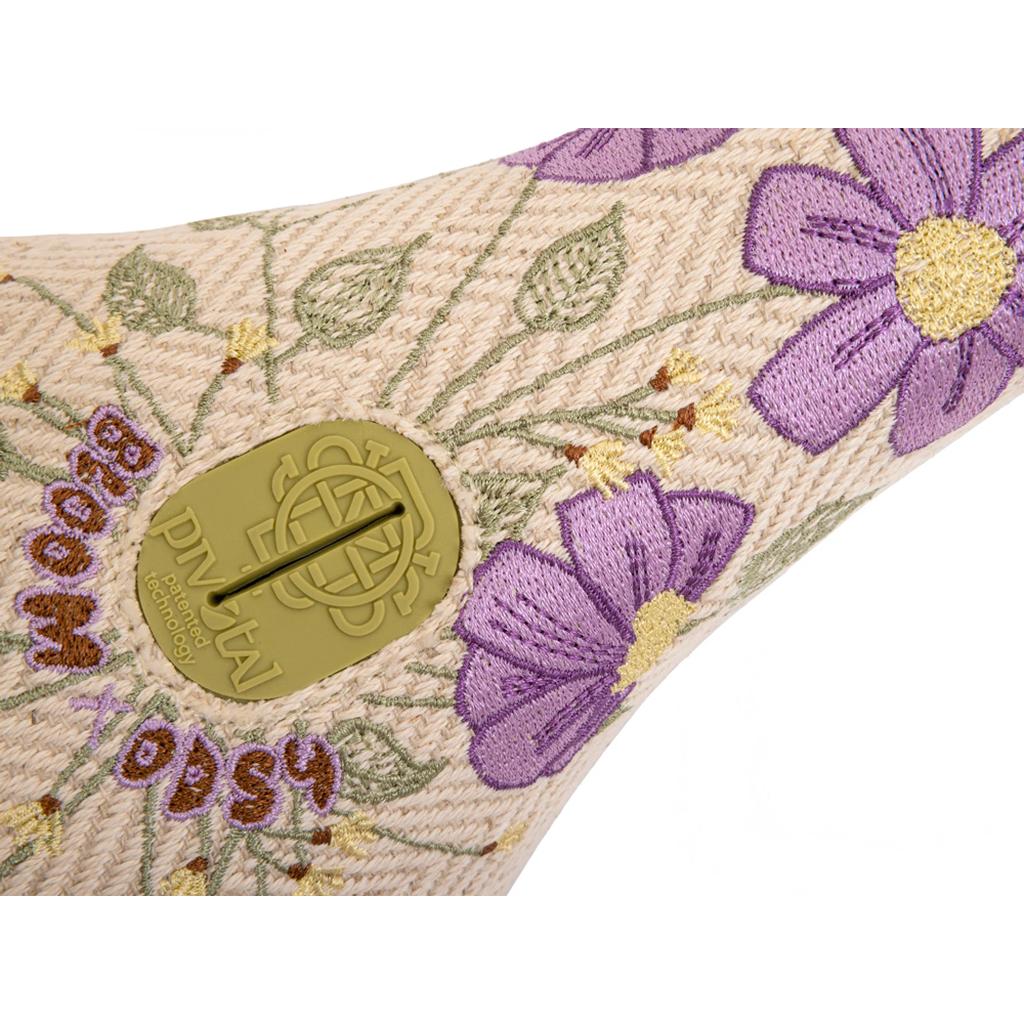 Close-up of the Odyssey Bloom V2 Pivotal Seat’s textured fabric, showcasing purple and yellow embroidered flowers, green leaves, and a tan plastic tag with a circular logo—offering stylish comfort for women riders.
