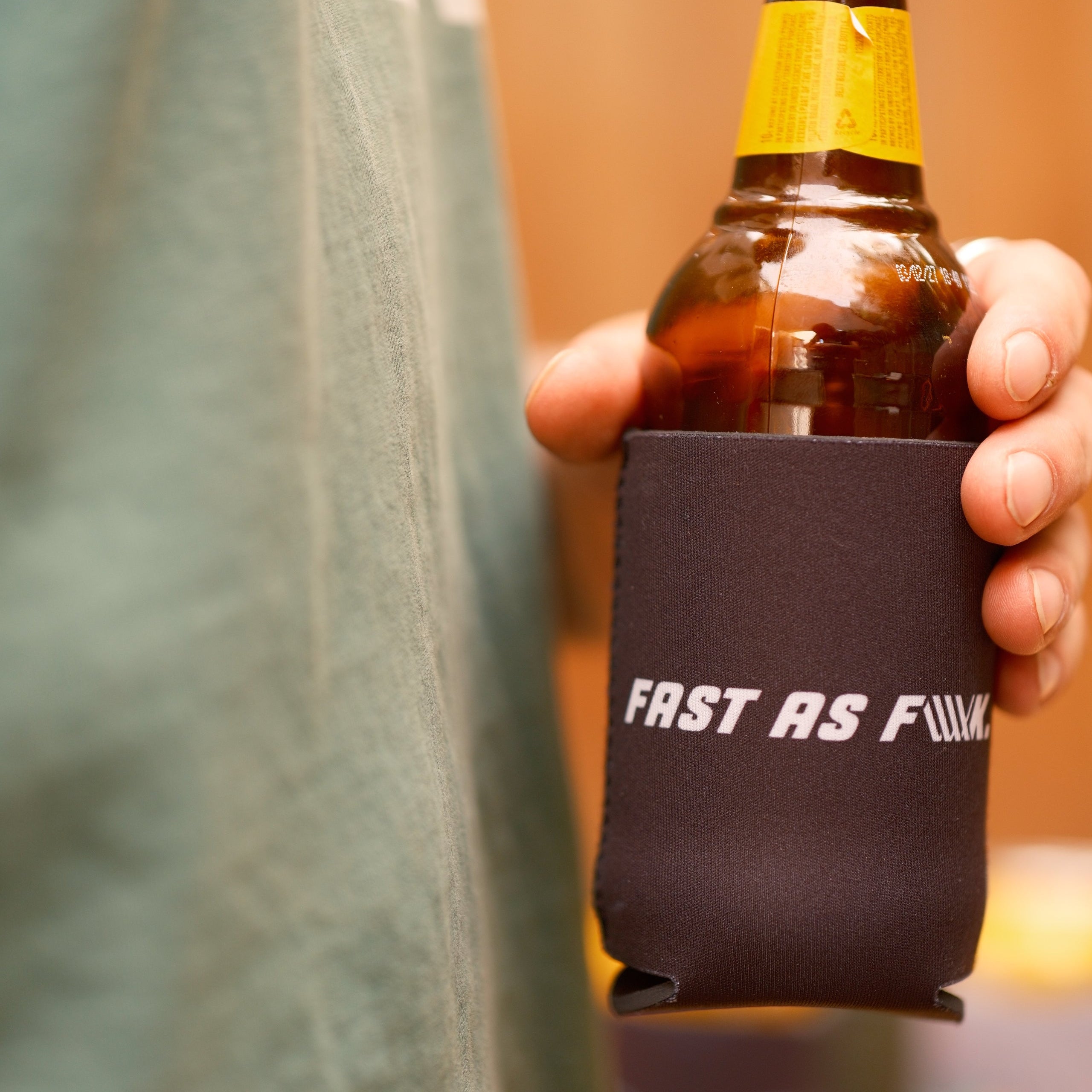 A person holds a brown glass bottle in a black LUXBMX Stubby Cooler / Fast As F-LUX, with part of the text obscured by their hand.