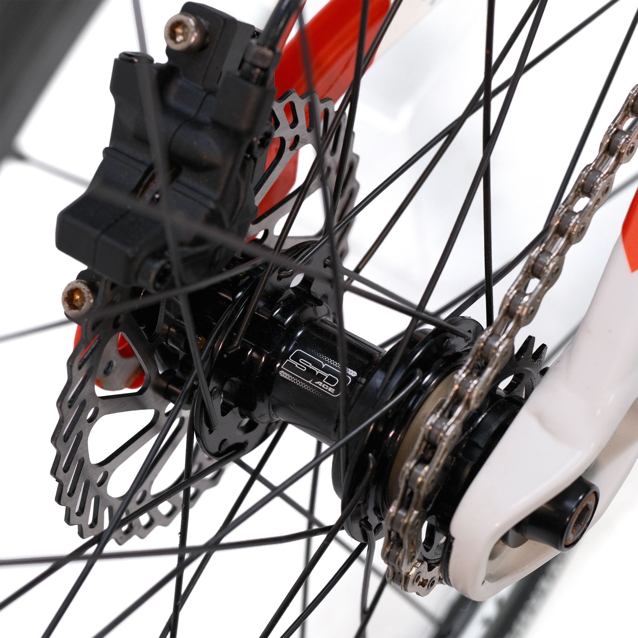 Close-up of the Stay Strong V5.1 Junior Race Bike rear wheel hub, highlighting the disc brake rotor, caliper, spokes, and chain on its lightweight red and white aluminium frame.