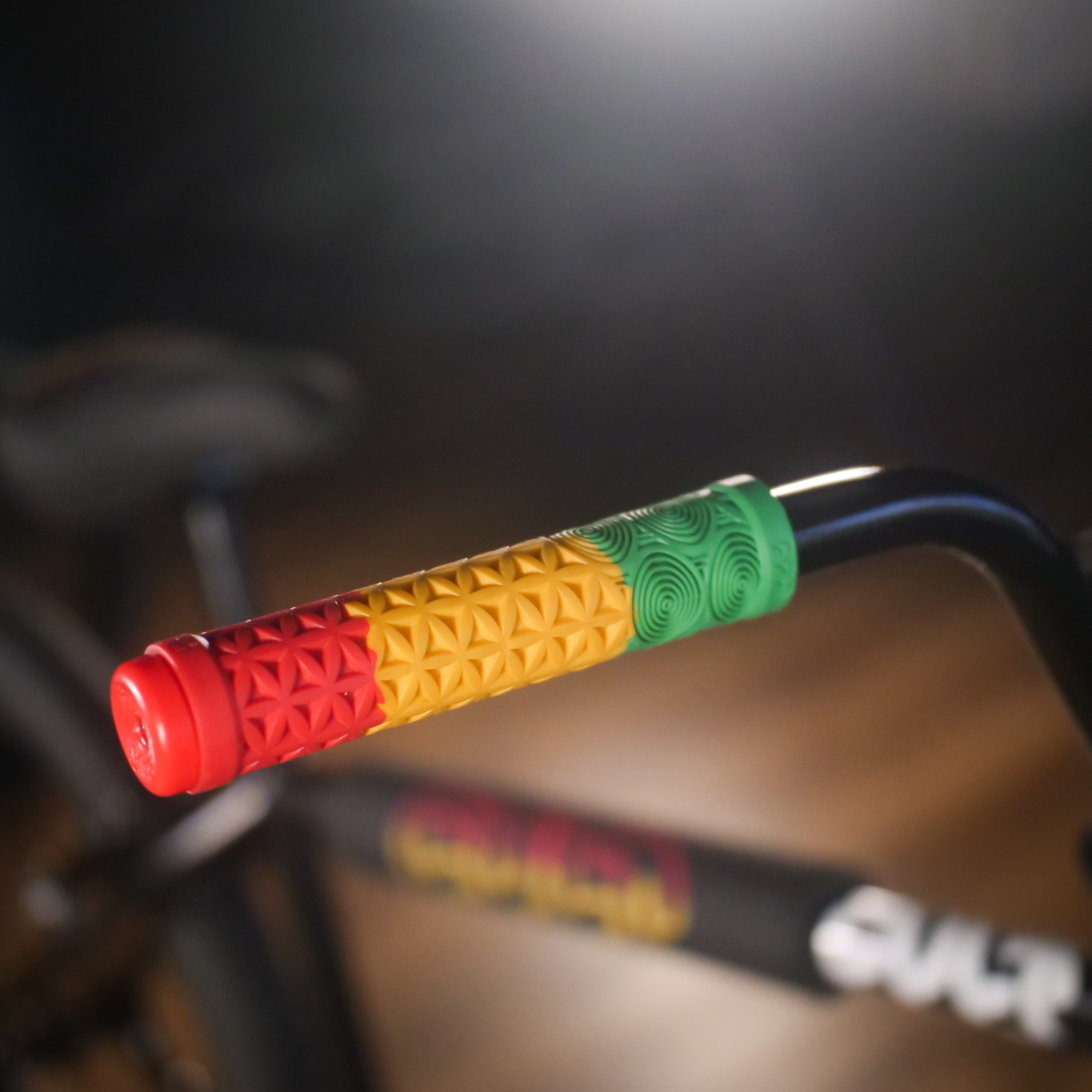 Close-up of the Cult Devotion 26 Inch Bike’s handlebar grip, showcasing red, yellow, and green textured sections with the Vans waffle tyres visible in the blurred background.