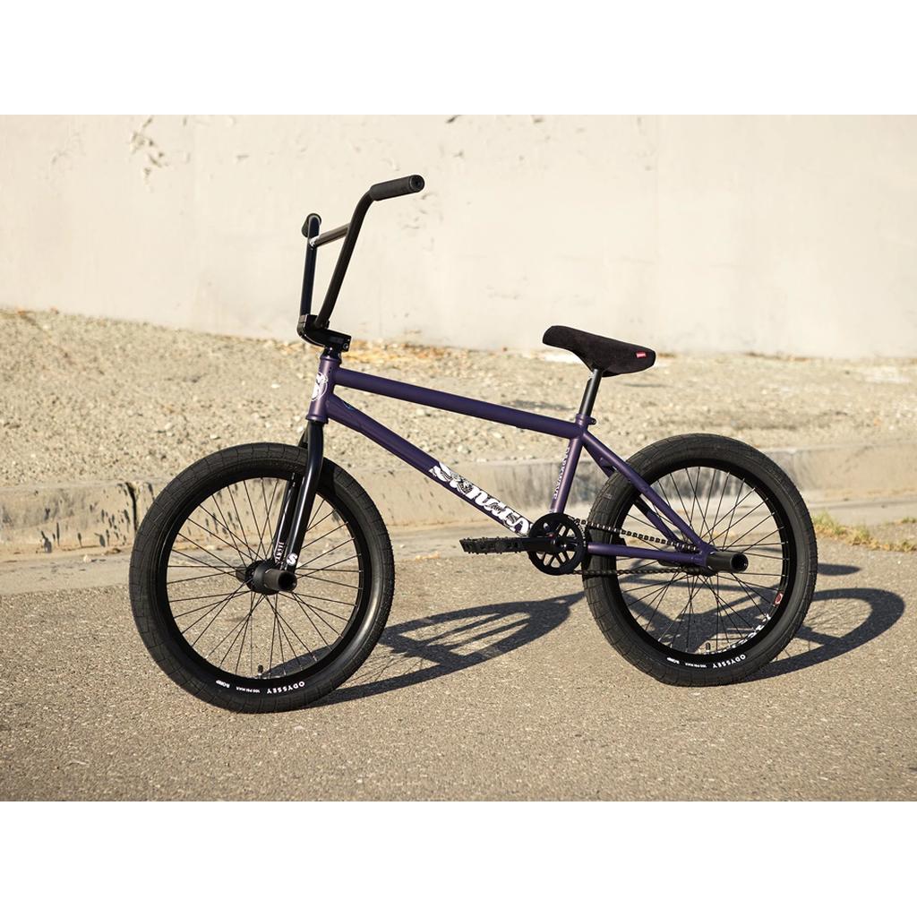 A Sunday Dark Wave Authentic 20 Inch Bike with a purple 100% Chromoly frame and black wheels and handlebars is parked on pavement near a concrete curb and gravel, highlighting its durable construction.