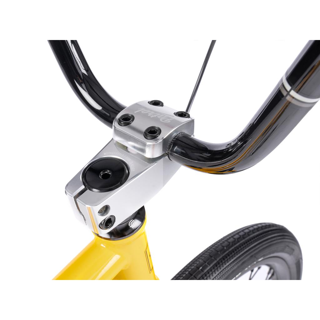 Close-up of a black BMX handlebar with a silver stem on the yellow Wethepeople Atlas 24 Inch Bike frame, showing part of the front tire and headset against a white background.