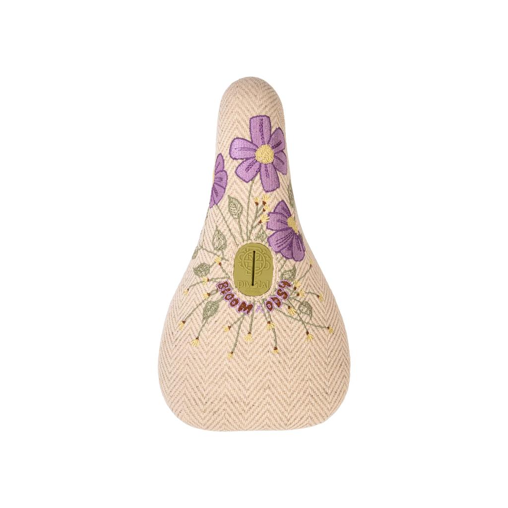 The Odyssey Bloom V2 Pivotal Seat features a beige woven texture, embroidered purple flowers, and a gold center emblem—ideal for women riders who value both stylish design and comfort.