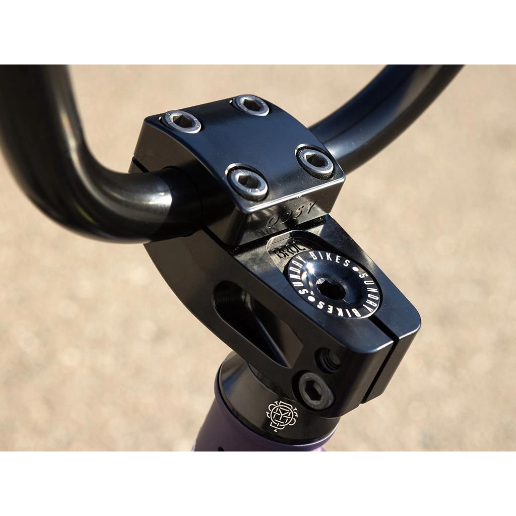 Close-up of a BMX bike stem clamping black handlebars, showing bolts and "Sunday Bikes" branding on the headset cap from the Sunday Dark Wave Authentic 20 Inch Bike.