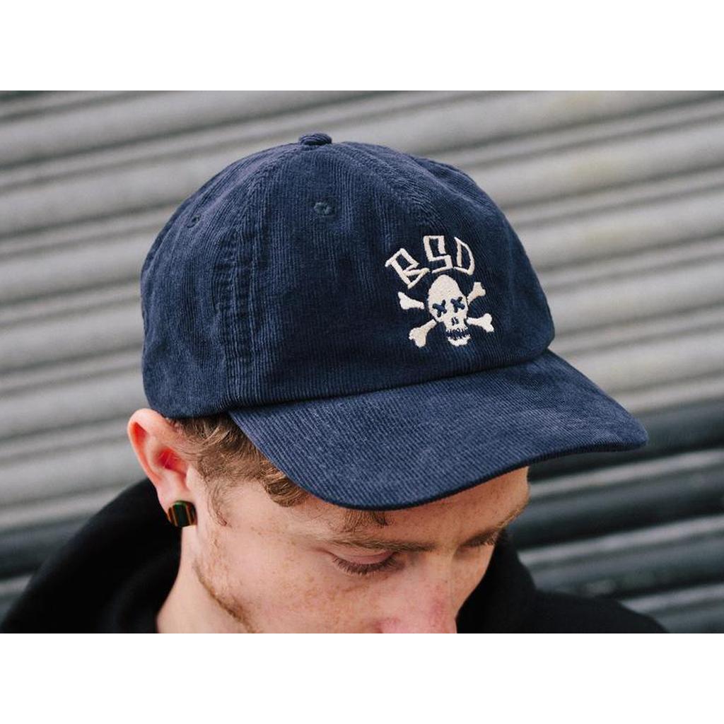 A person wearing the BSD More Speed Cap, featuring a blue color and skull and crossbones logo, stands in front of a metal shutter.