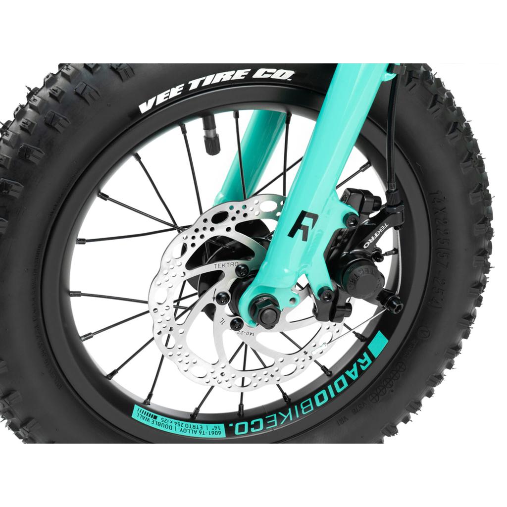 Close-up of the Radio Zuma 14 Inch Bike’s teal front wheel, highlighting a Vee Tire Co. tire, Tektro disc brake, and spoke details—ideal features for a lightweight kids’ mountain bike.