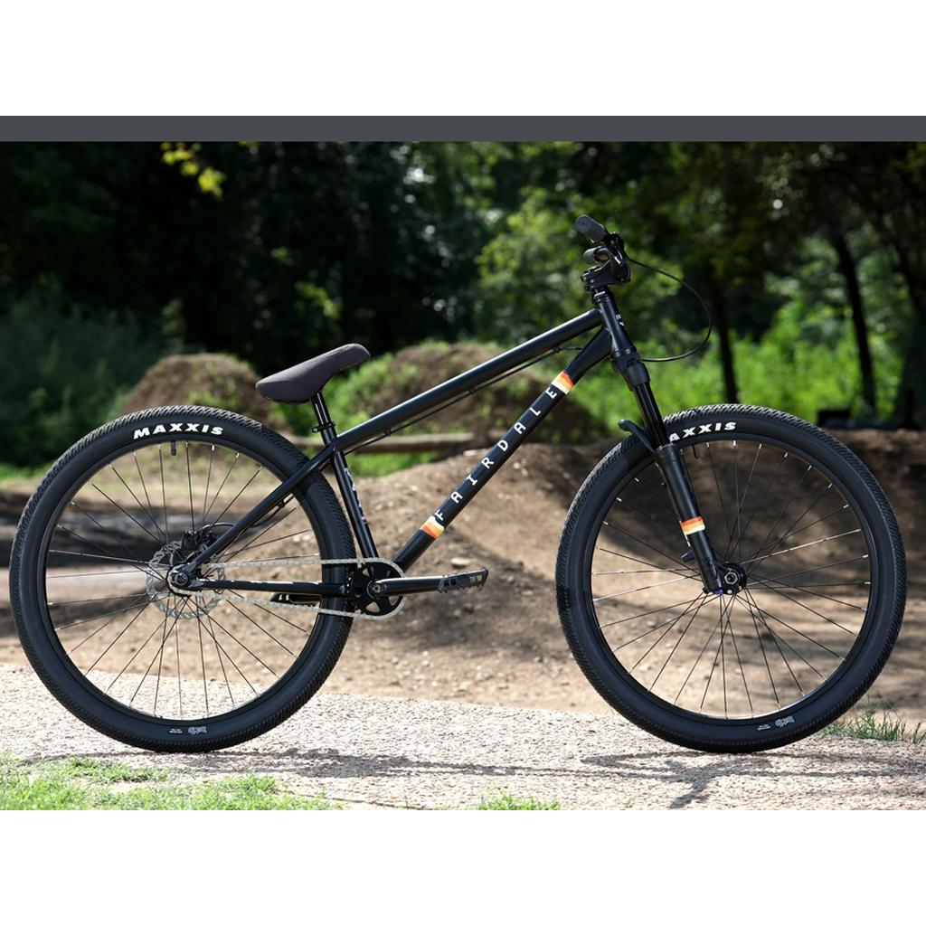 A Fairdale Hareraiser FX 26 Inch Bike with a black frame and Maxxis tires is parked on a gravel path, surrounded by greenery and dirt mounds.