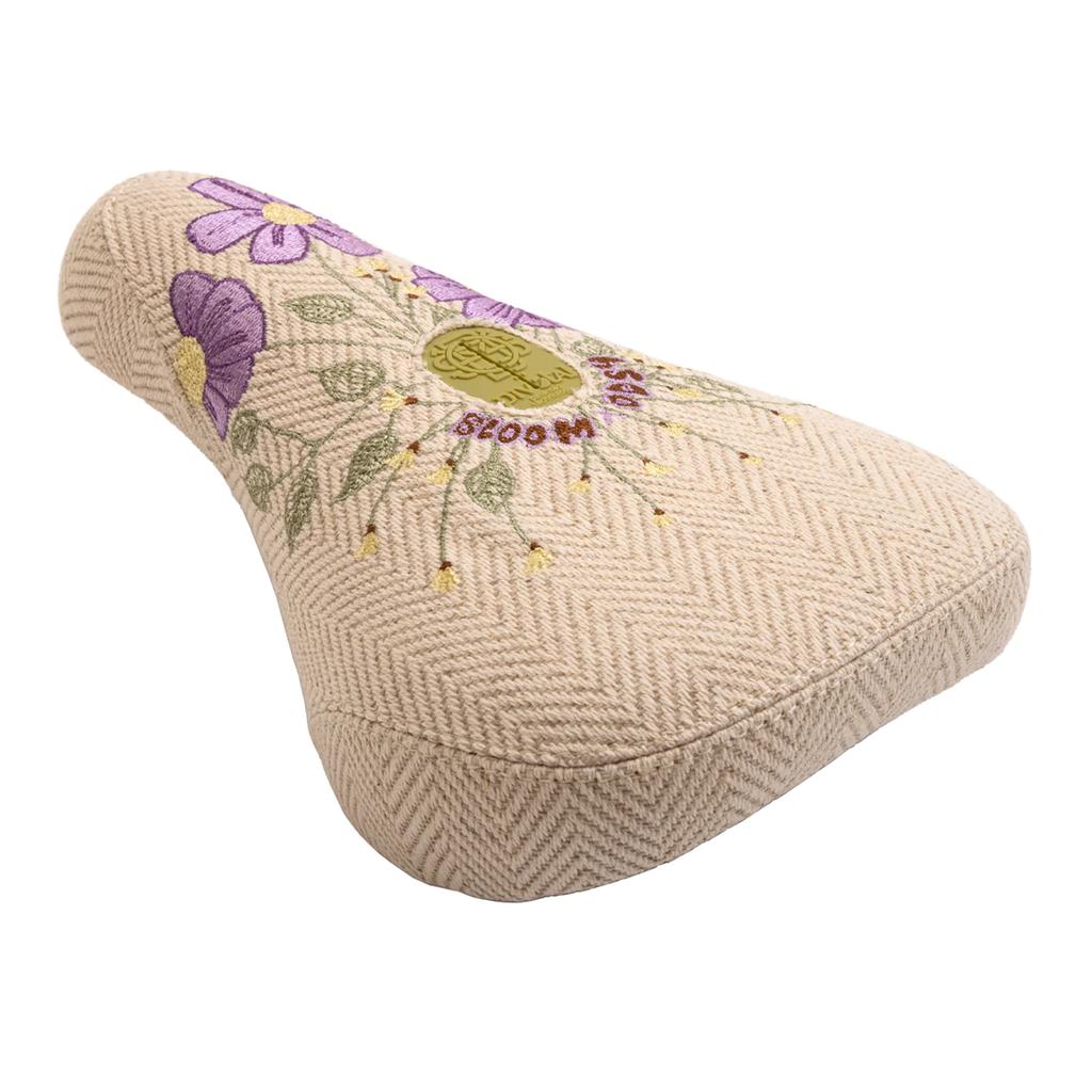 The Odyssey Bloom V2 Pivotal Seat is beige with a herringbone pattern, embroidered purple flowers and green leaves, plus "BLOOM DASH" in red—stylish and ideal for women BMX riders.
