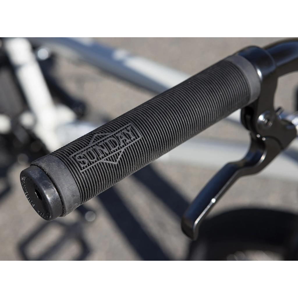 Close-up of a black bike handlebar grip featuring the "SUNDAY" logo and paired with an Odyssey Springfield brake lever, set against a blurred outdoor background—ideal for riders who want to customize their Sunday Blueprint 20 inch Bike or upgrade any entry-level BMX setup.