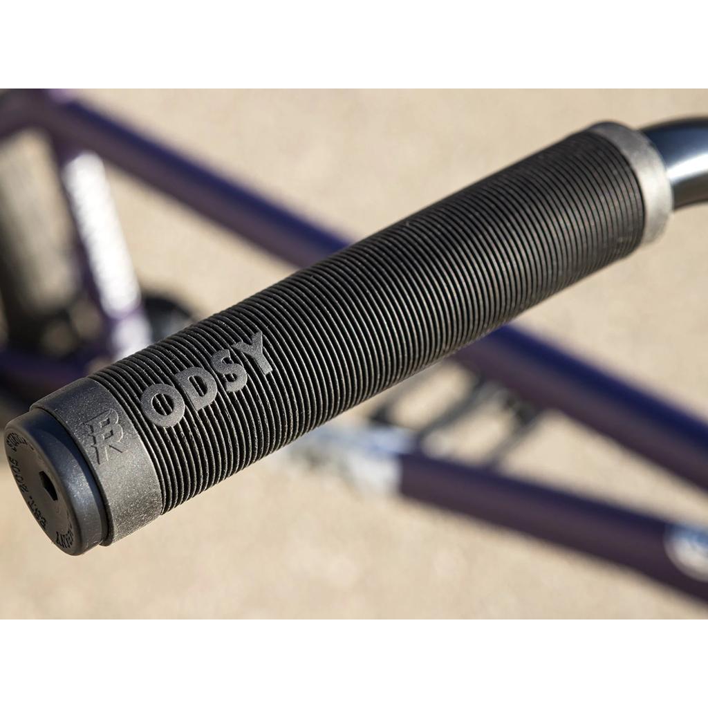 Close-up of a black handlebar grip with "ODSY" and logo embossed; the 100% Chromoly frame of the Sunday Dark Wave Authentic 20 Inch Bike is blurred in the background.