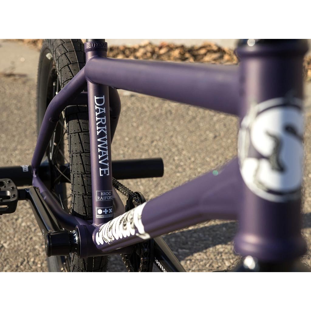 Close-up of a Sunday Dark Wave Authentic 20 Inch Bike with "DARKWAVE" on the tube, highlighting its 100% Chromoly frame, rear wheel, and pedal. This is Broc Raiford’s signature BMX model on a paved surface.
