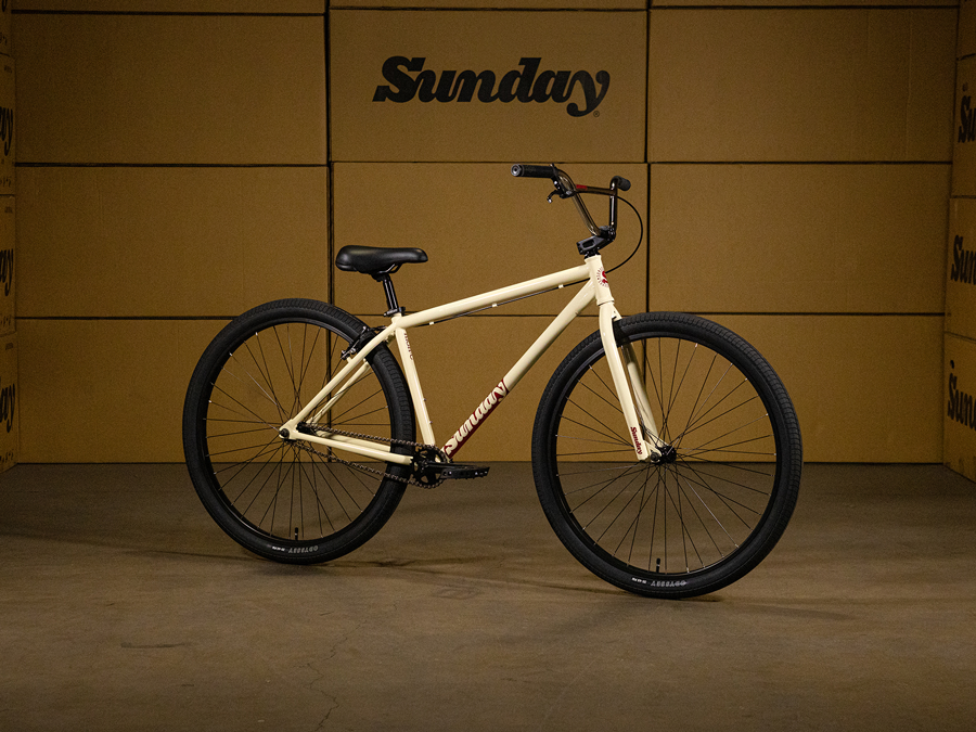 The Sunday High C 29 Inch Bike—a cream-colored BMX-style bicycle with black tires—stands indoors before stacked cardboard boxes marked "Sunday." This 29" cruiser boasts a 100% chromoly frame for both superior strength and timeless style.