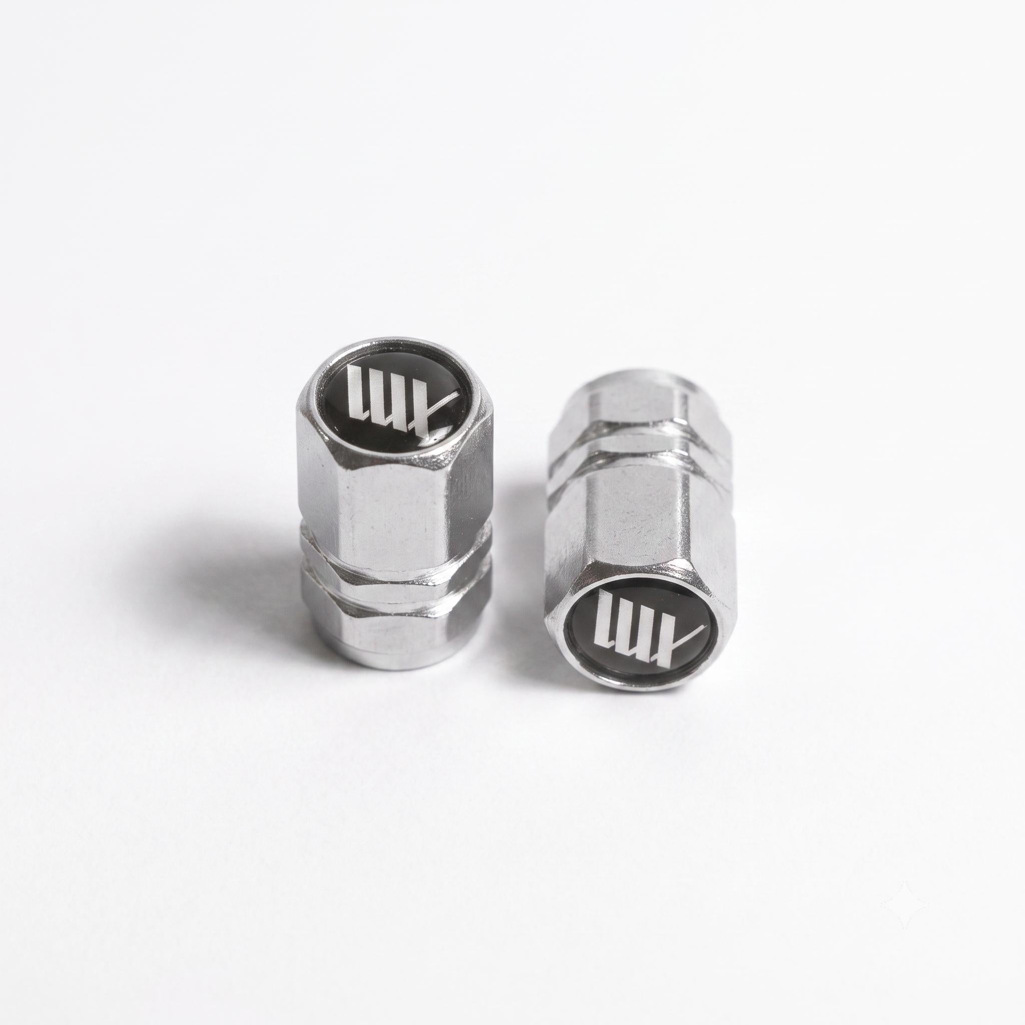 Two LUXBMX Hex Logo Alloy Valve Caps stand upright on a white surface, each resembling machined metal and featuring the LUX logo on top.