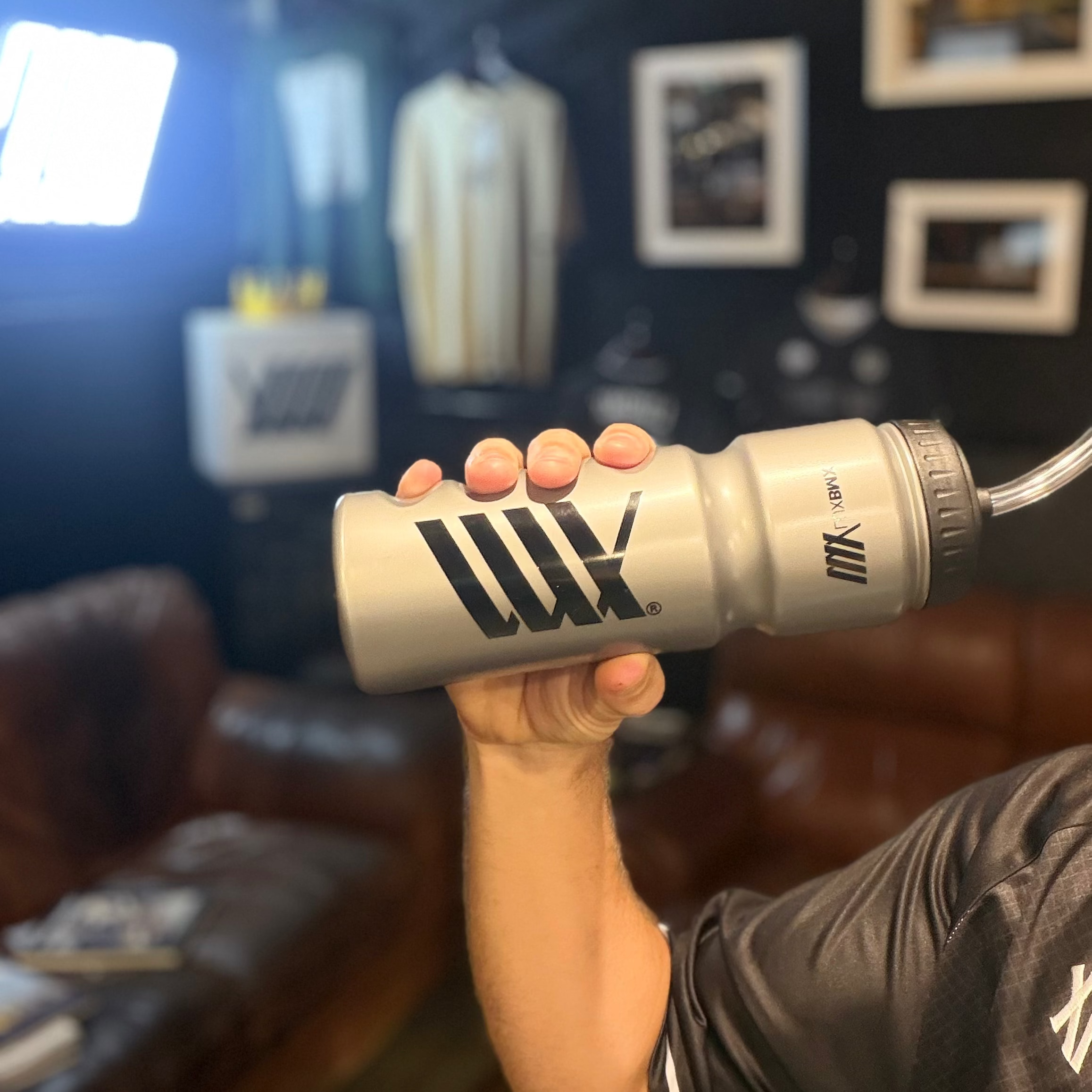 A person holds a gray LUXBMX Race Straw Water Bottle 750ml, made from BPA-free plastic, in a room featuring framed pictures, a brown couch, and hanging shirts in the background.