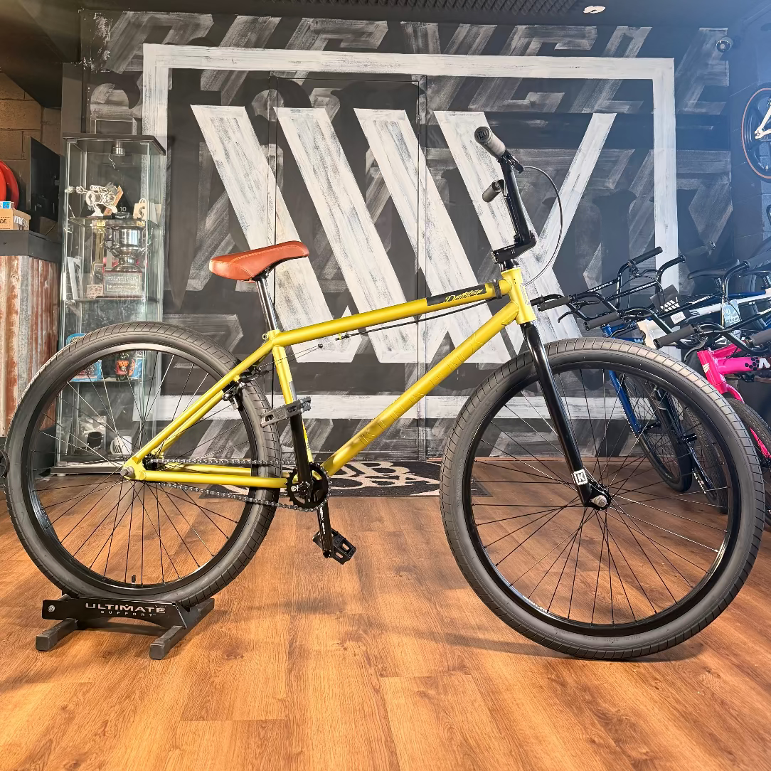 A Kink Drifter 26 inch Bike (2027), featuring a yellow frame and brown seat, is displayed on a stand in a bike shop with wooden floors and black-and-white wall art in the background.