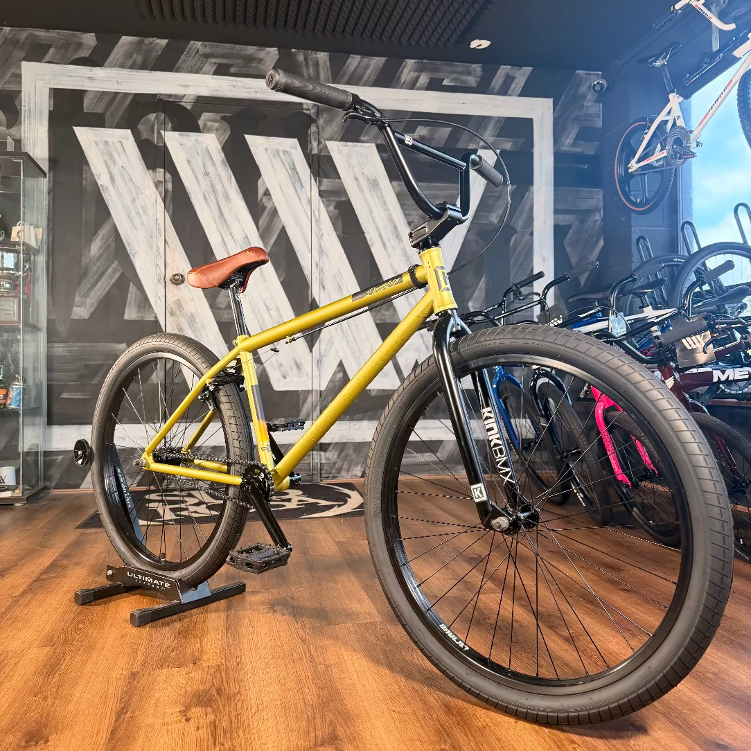 The Kink Drifter 26 inch Bike (2027), featuring a yellow frame and brown seat, is shown indoors on a wooden floor with other bicycles and gear in the background.