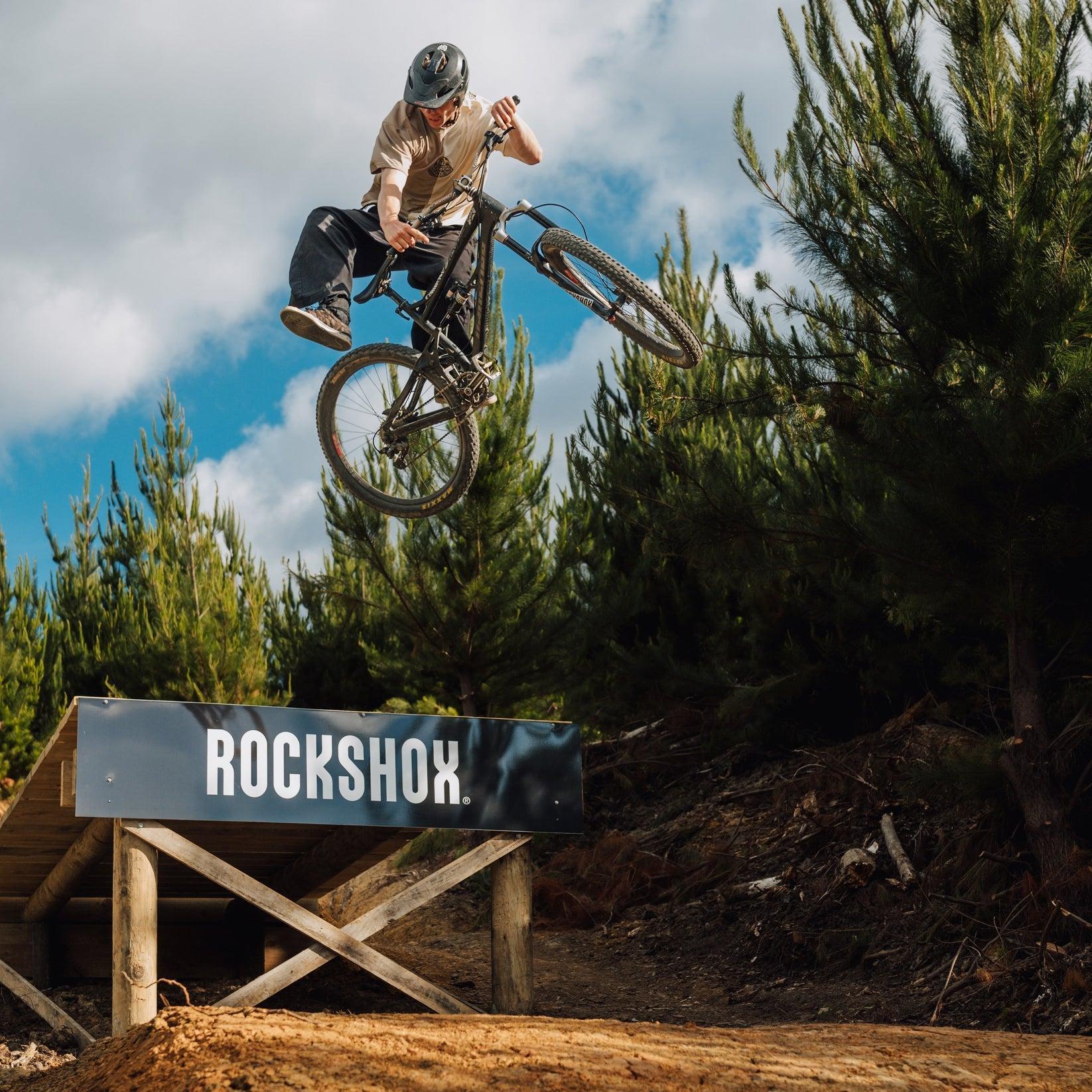 A person wearing a helmet and the Buzzed Riders Split Tee catches air on a mountain bike over a wooden ramp branded with "RockShox," surrounded by trees and partly cloudy skies—an action-sport moment that's sure to spark conversation.