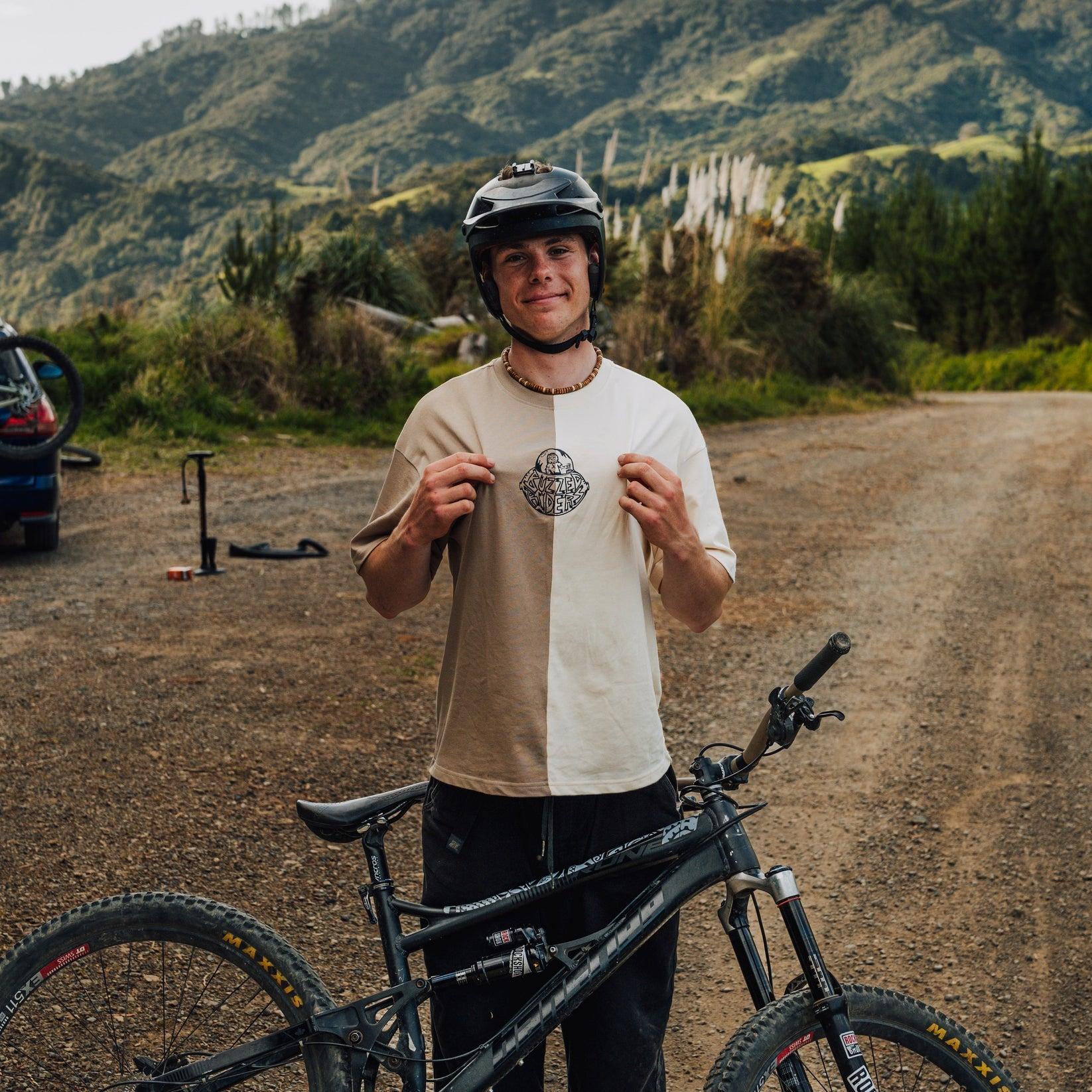 A young person in a Buzzed Riders Split Tee and helmet stands beside a mountain bike, proudly holding up a medal on a dirt road with mountains in the background—a dynamic action sport moment that’s sure to spark conversation.