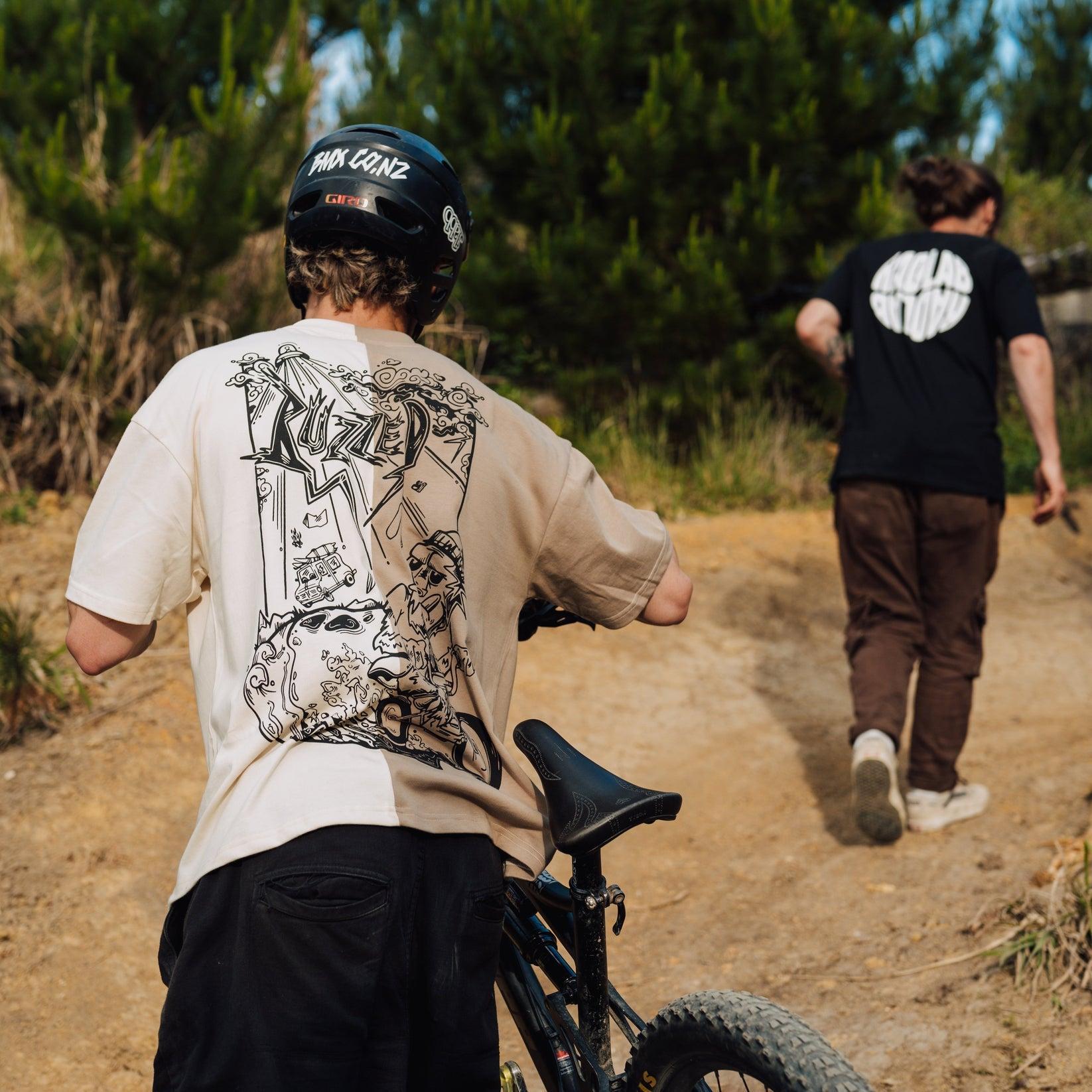Two people make their way along a dirt trail; one sports the Buzzed Riders Split Tee while walking a bike uphill, the other walks ahead. Both are outfitted in casual clothes and helmets—a perfect action sport scene set against trees and grass.