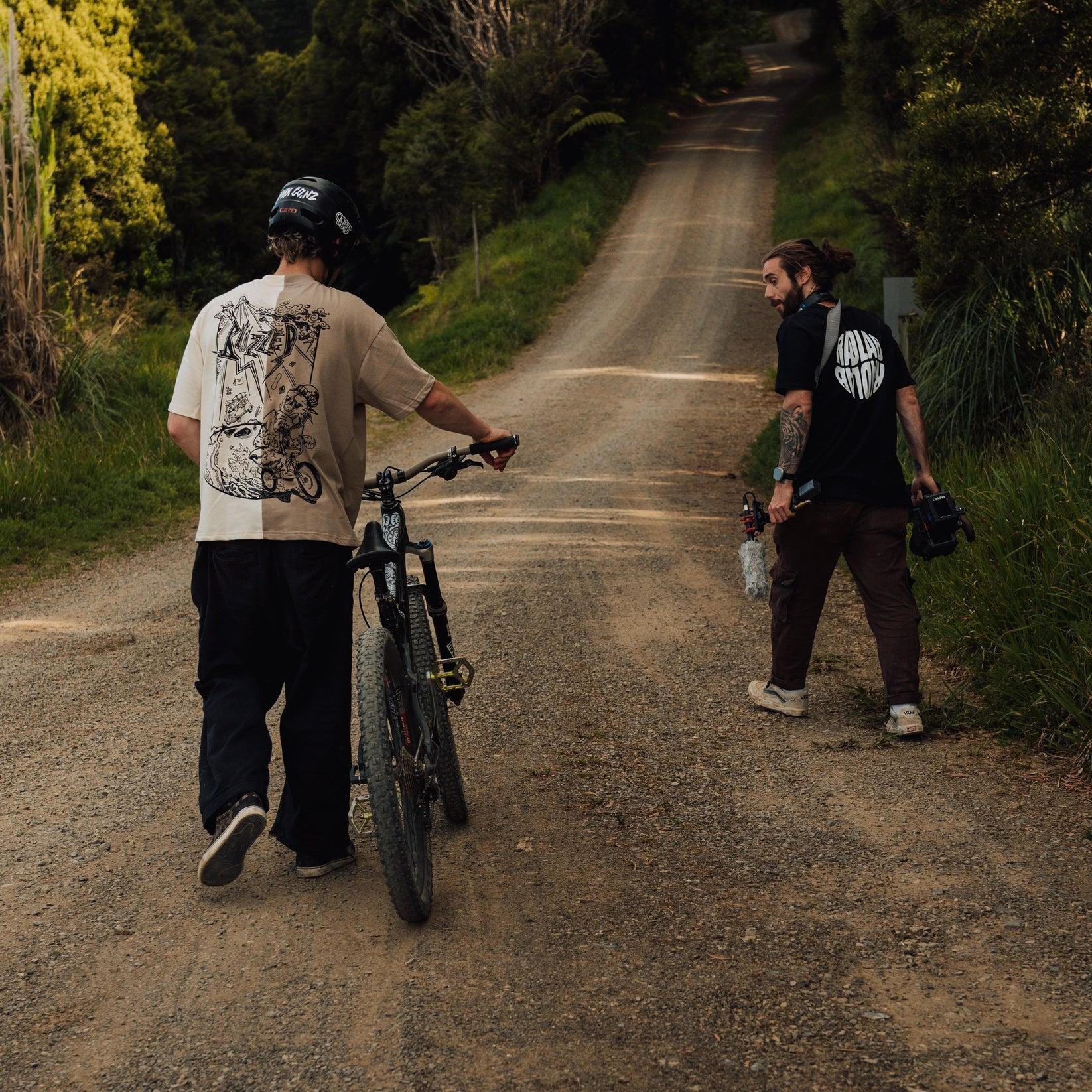 Two people walk up a dirt road in a forested area; one, dressed in the Buzzed Riders Split Tee, pushes a mountain bike—clearly ready for some action sport—while the other carries camera equipment and a drink.