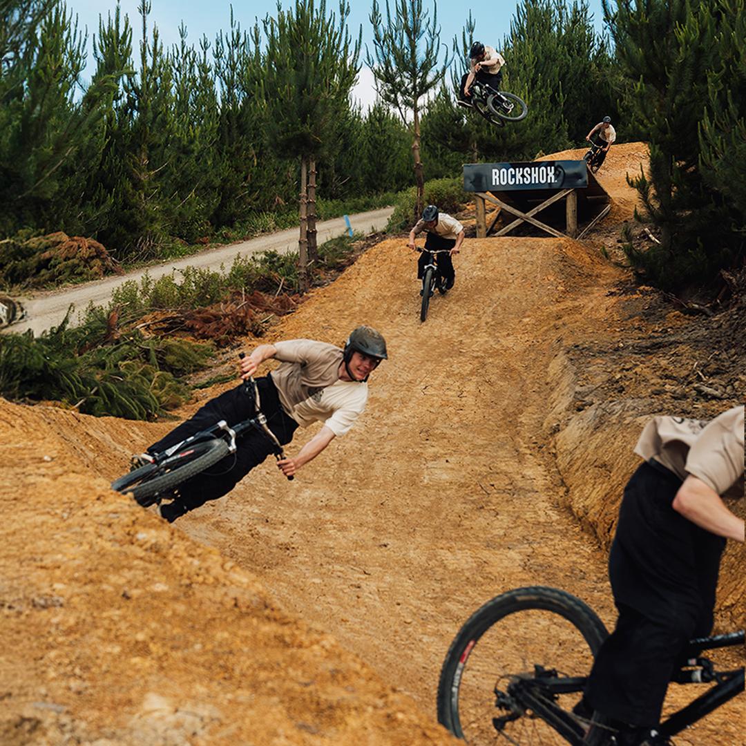 Four cyclists, each sporting the Buzzed Riders Split Tee, ride mountain bikes on a dirt trail with jumps beneath towering pine trees. In the background, a ramp marked "RockShox" hints at high-flying action—making this vibrant scene a true conversation starter.