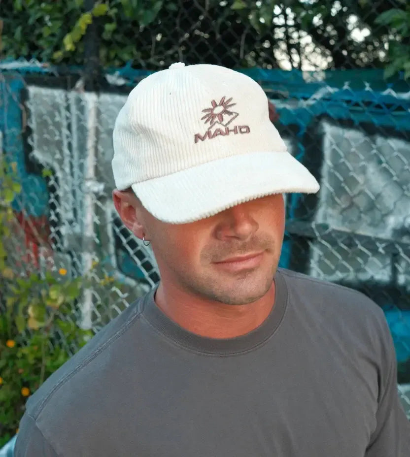 A man in the Mahd Botanical Corduroy Hat and a gray tee stands before a chain-link fence, with lush greenery in the background.