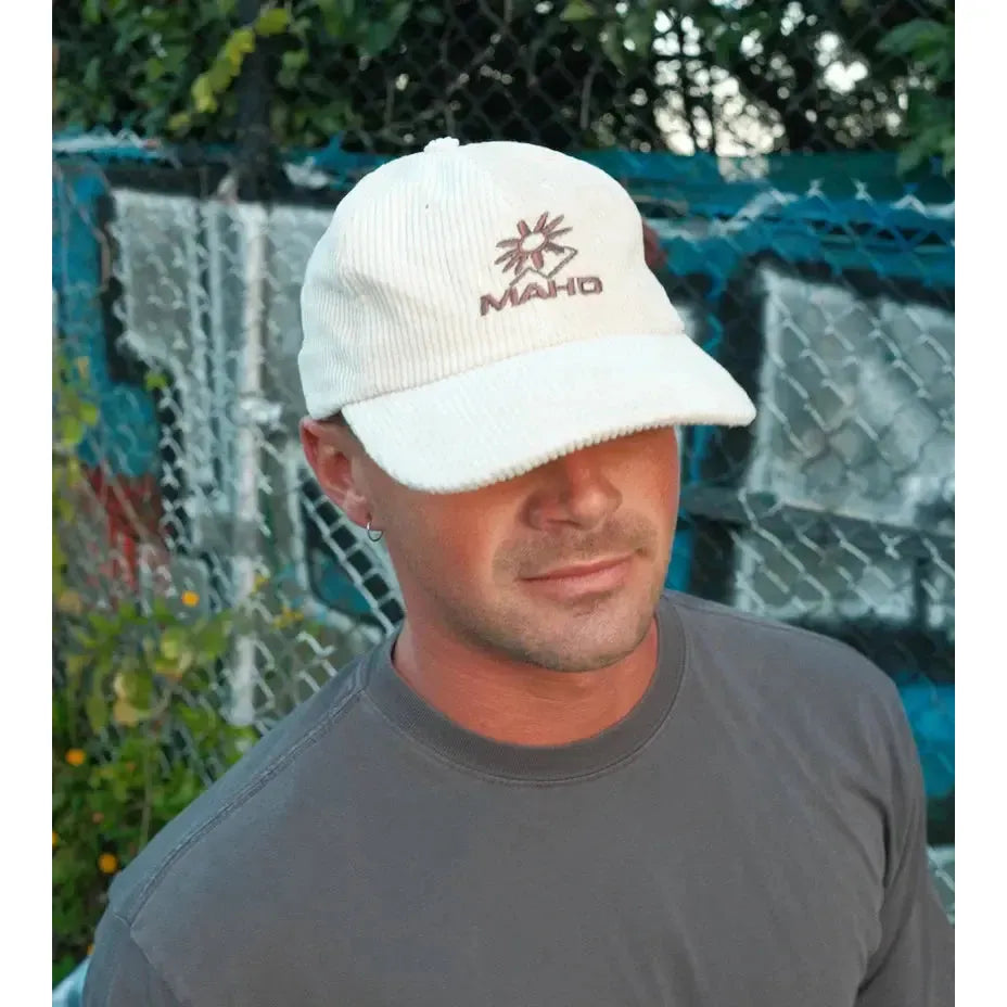 A man in the Mahd Botanical Corduroy Hat and a gray tee stands before a chain-link fence, with lush greenery in the background.