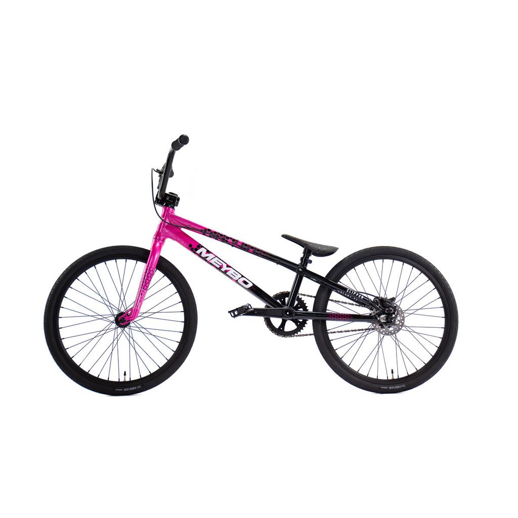 The Meybo 2026 Clipper Expert XL Bike, shown in pink and black with a straight top tube, knobby tires, and black handlebars, is displayed in side view. Ideal as an entry-level BMX race bike for new riders.