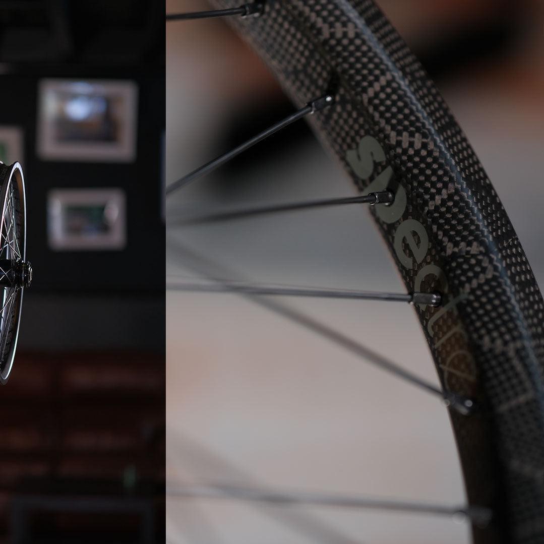 Close-up of a Spectre NME X Onyx Elite 20 Inch Disc Wheel Set (20mm/15mm) with black spokes and a carbon fiber rim. The brand name is partially visible, with blurred picture frames in the background.