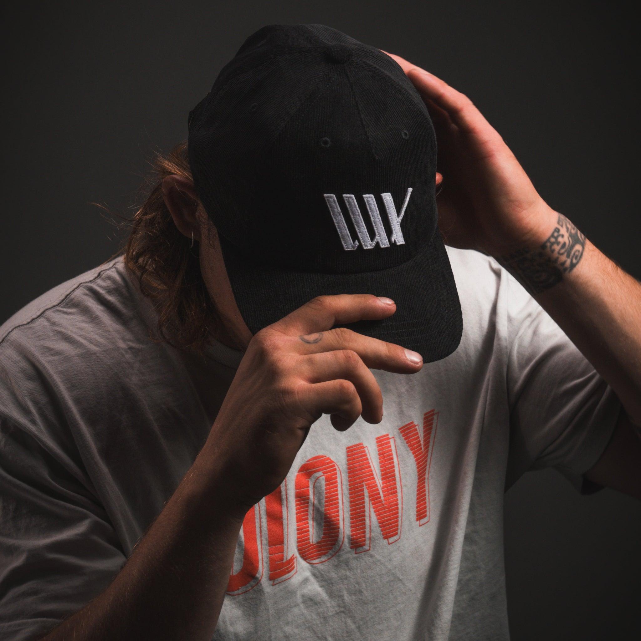 A person in a LUXBMX Outfield Cap Black with an embroidered LUX logo, paired with a white T-shirt featuring red text, tilts their head down and covers part of their face with one hand.