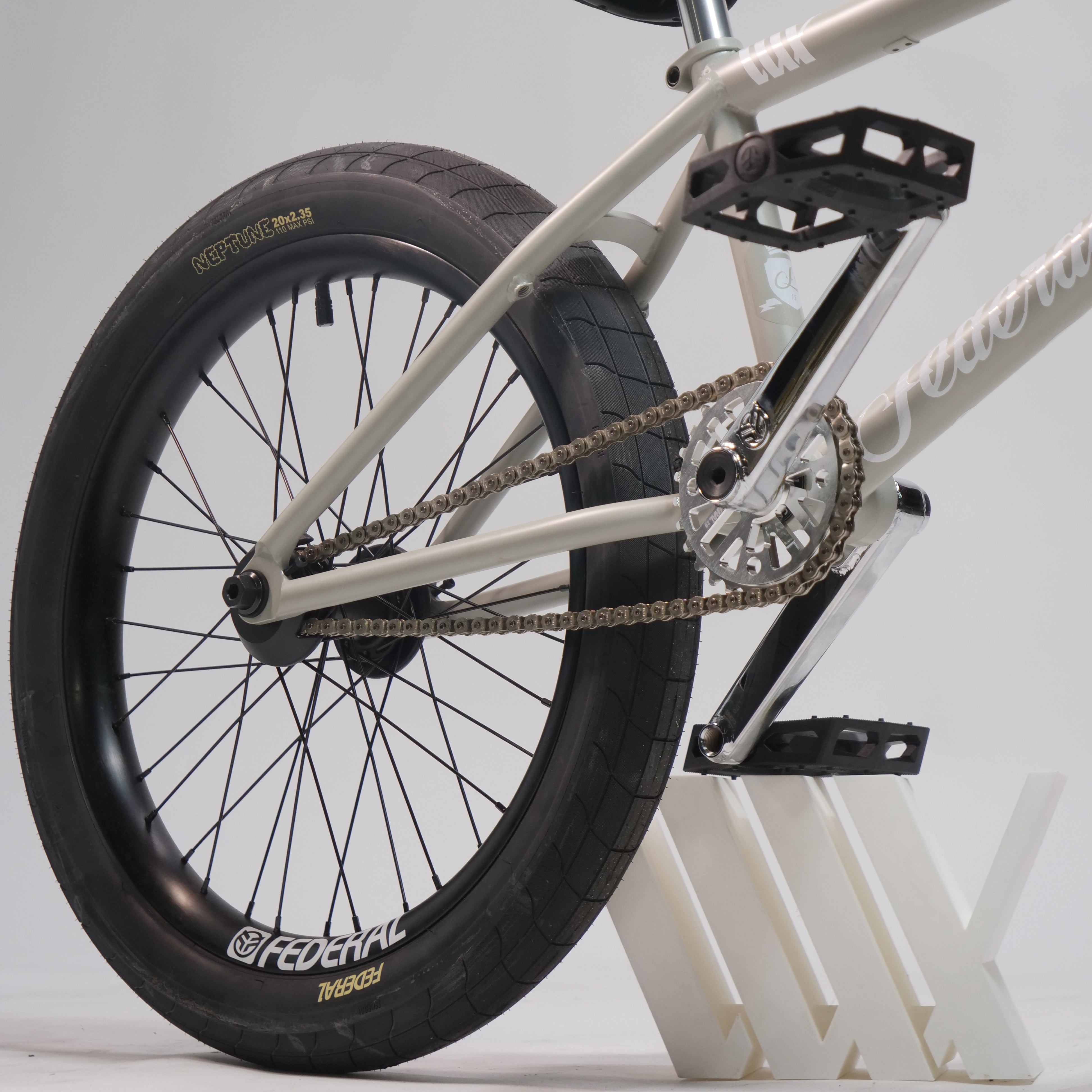 Close-up of the rear wheel, chain, pedals, and frame of the Federal Lacey x Chrome Custom 20 Inch Bike on a white stand against a plain background.