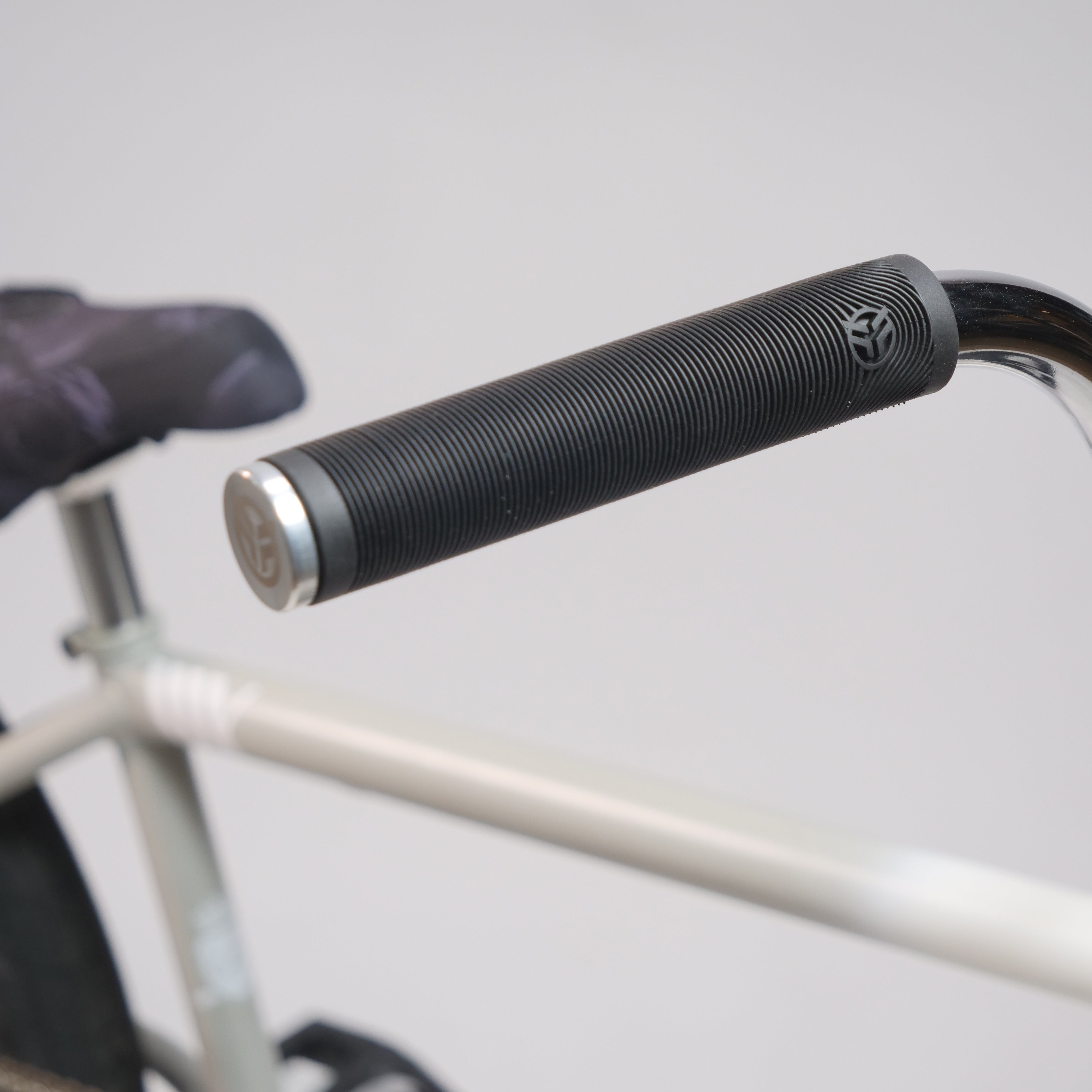 Close-up of a Federal Lacey x Chrome Custom 20 Inch Bike handlebar with a black textured grip and metallic end cap, plus visible frame, seat, and high-performance Federal Vice 2 Cranks in the background.