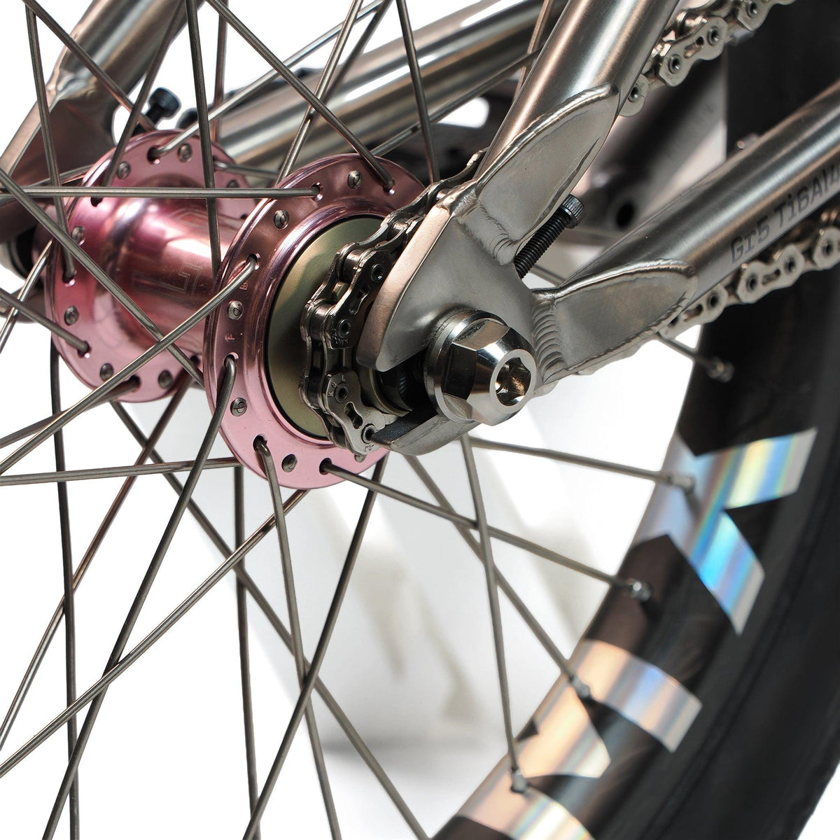 Close-up of the ION BMX Ti18 Custom Titanium 18 Inch Bike’s rear wheel hub, showing metallic spokes, chain, silver frame, detailed mechanical parts, and a tire with reflective accents on this lightweight freestyle BMX.
