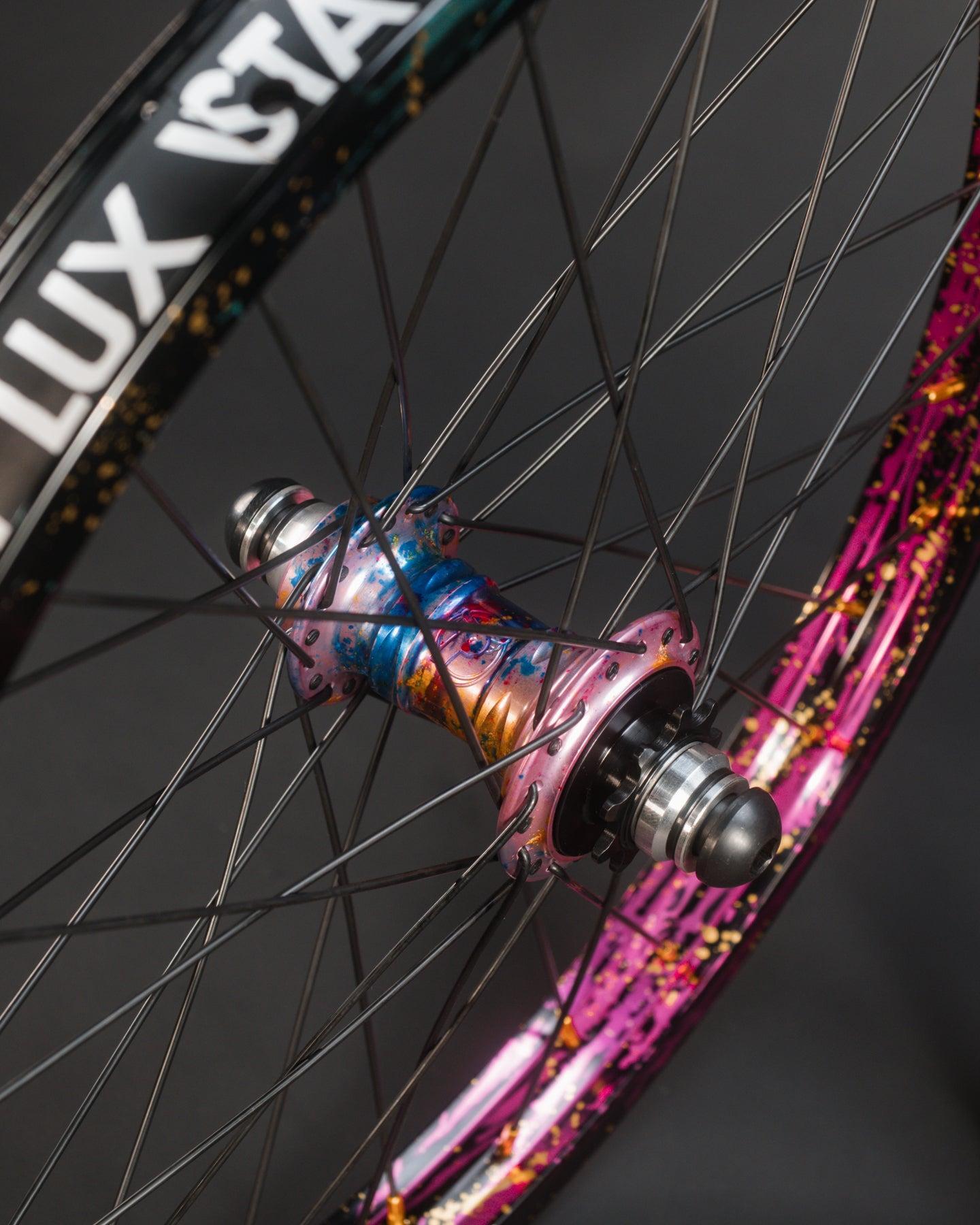Close-up of the Profile x Odyssey Near Earth Objects Wheel Set (Limited Edition) featuring black spokes, rims with "LUX" text, and a vibrant splatter paint design.