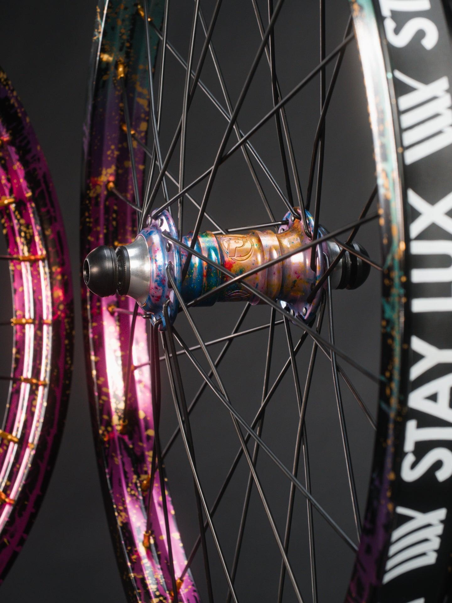 Close-up of a Profile x Odyssey Near Earth Objects Wheel Set (Limited Edition) featuring black spokes, a metallic multicolor BMX hub, and an Odyssey Hazard 40th Anniversary Rim labeled "STAY LUX." Another similar wheel appears in the background.