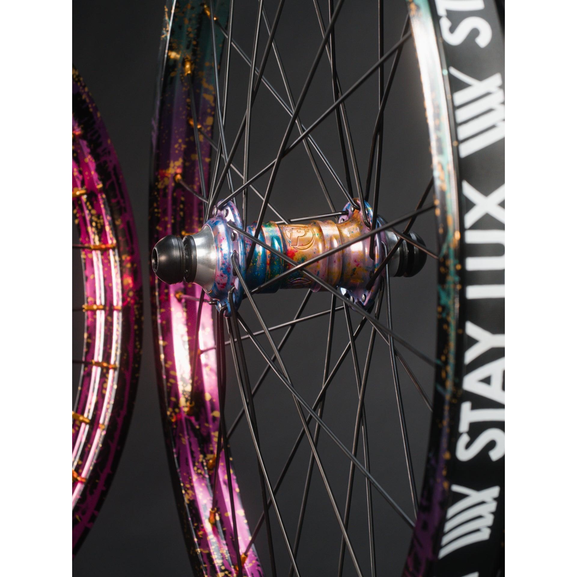 Close-up of a Profile x Odyssey Near Earth Objects Wheel Set (Limited Edition) featuring black spokes, a metallic multicolor BMX hub, and an Odyssey Hazard 40th Anniversary Rim labeled "STAY LUX." Another similar wheel appears in the background.