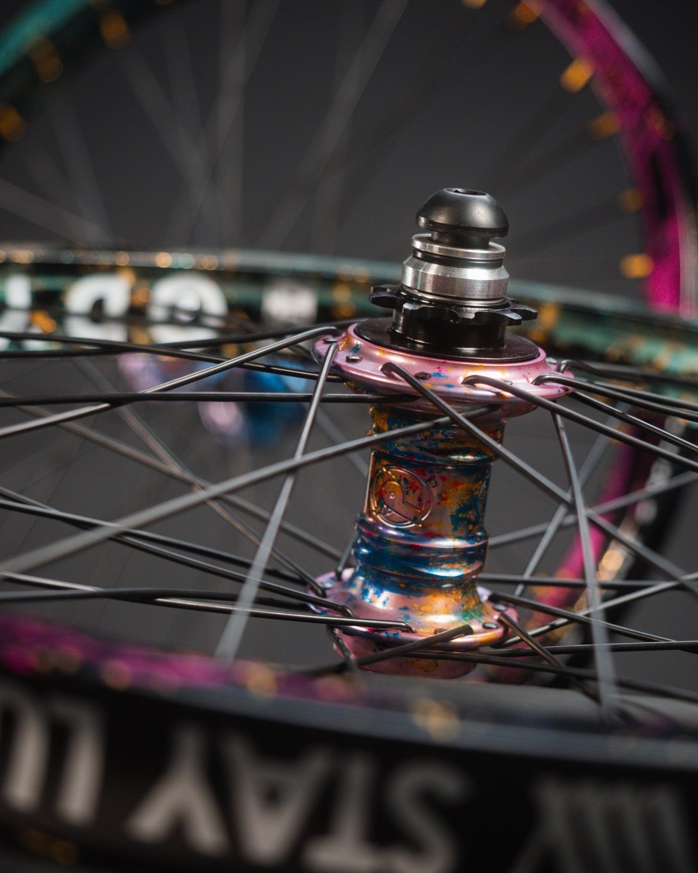 Close-up of the Profile x Odyssey Near Earth Objects Wheel Set (Limited Edition), featuring a vibrant BMX hub, Odyssey Hazard 40th Anniversary Rim, black spokes, and metallic finish. Another wheel appears blurred in the background.