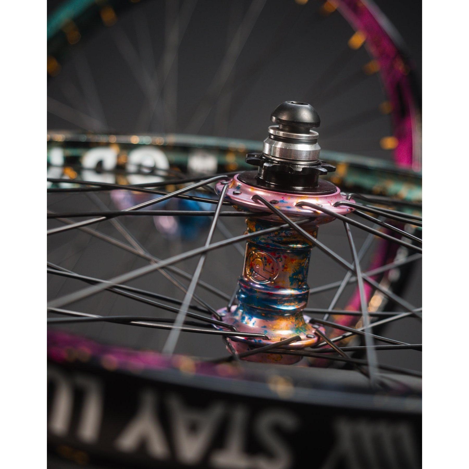 Close-up of the Profile x Odyssey Near Earth Objects Wheel Set (Limited Edition), featuring a vibrant BMX hub, Odyssey Hazard 40th Anniversary Rim, black spokes, and metallic finish. Another wheel appears blurred in the background.