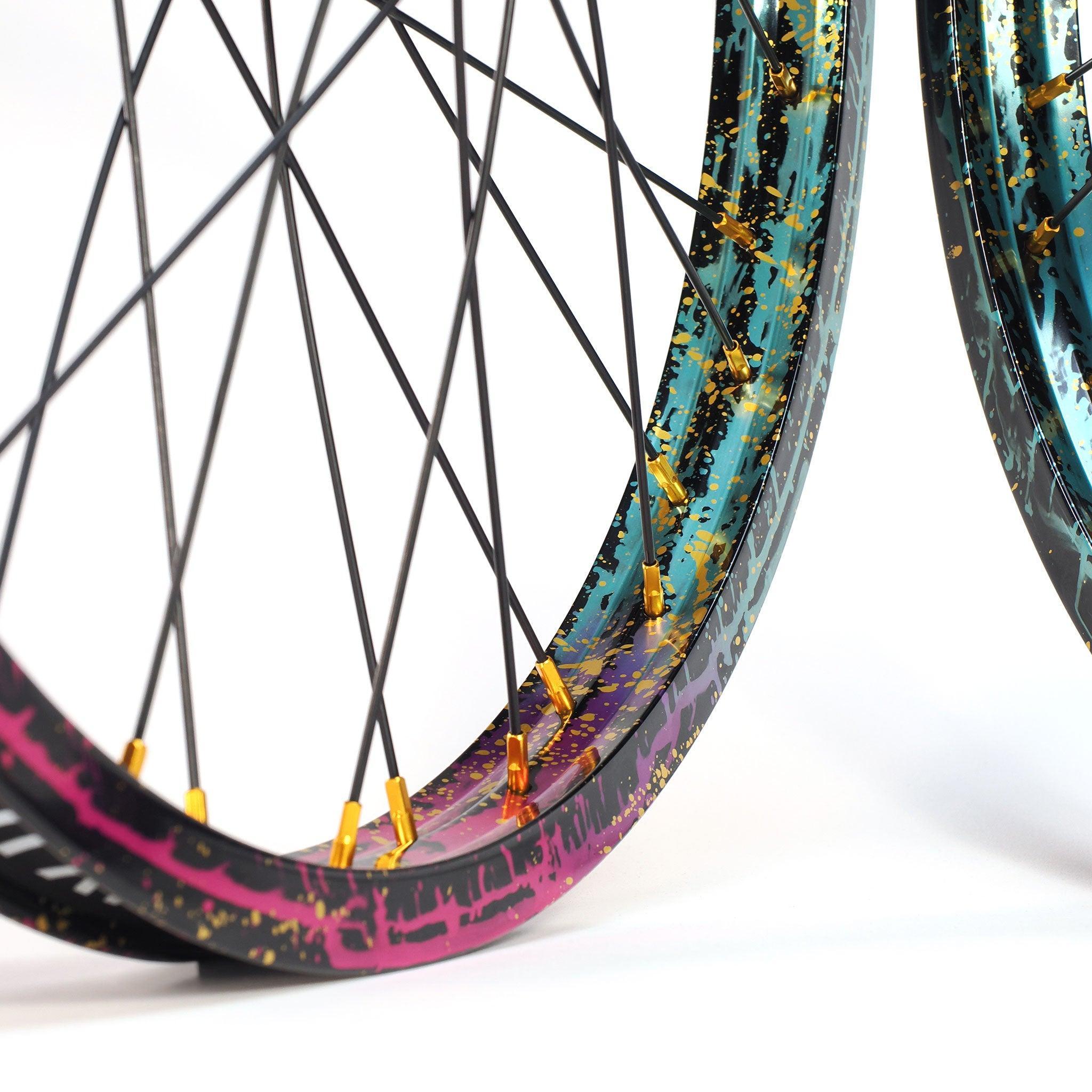 Close-up of two Profile x Odyssey Near Earth Objects Wheel Set (Limited Edition) bicycle wheels, featuring colorful, splattered paint on the rims and gold spoke nipples against a white background.