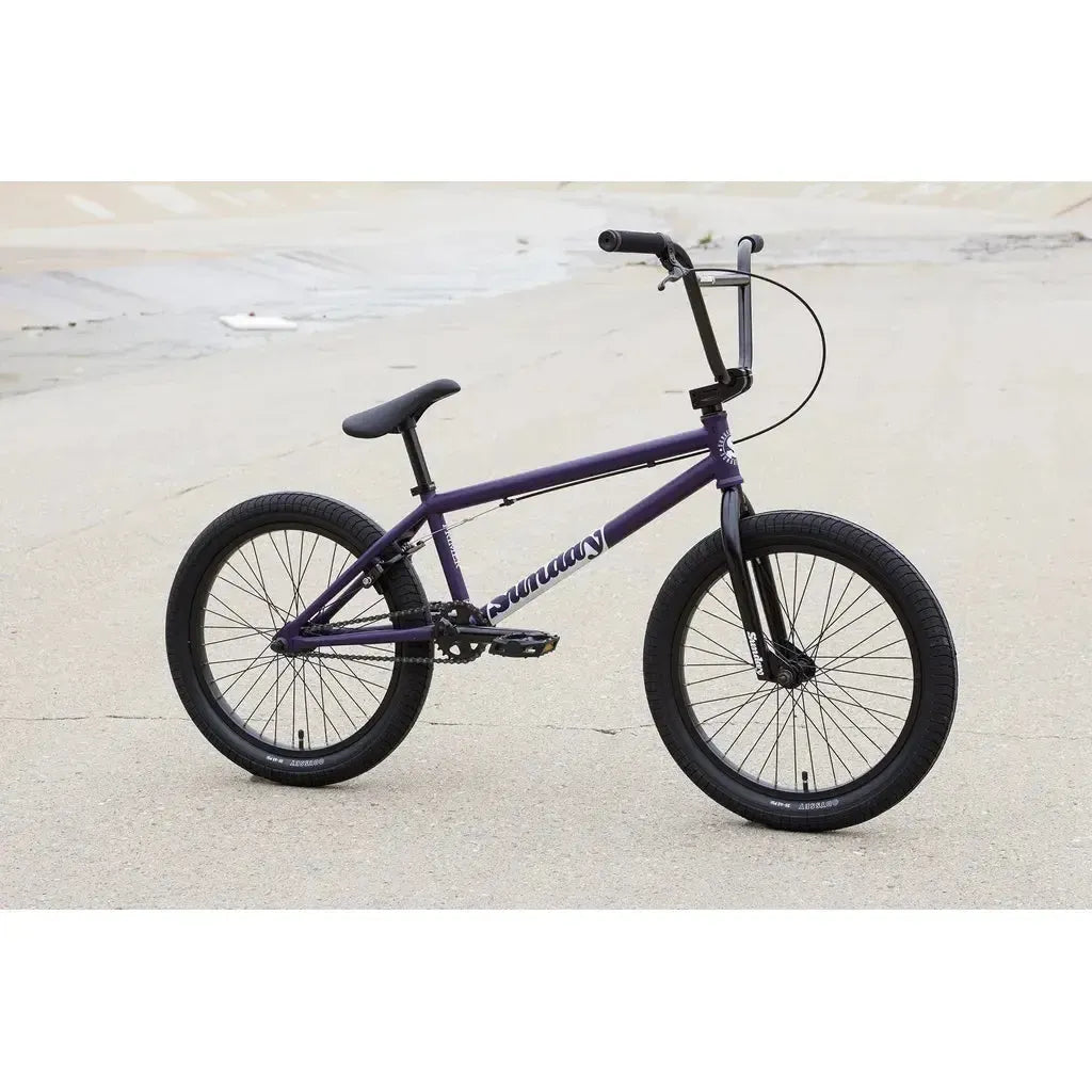 A Sunday Primer 20 Inch Bike in purple with black tires and seat is parked on a concrete surface near a sloped drainage area.