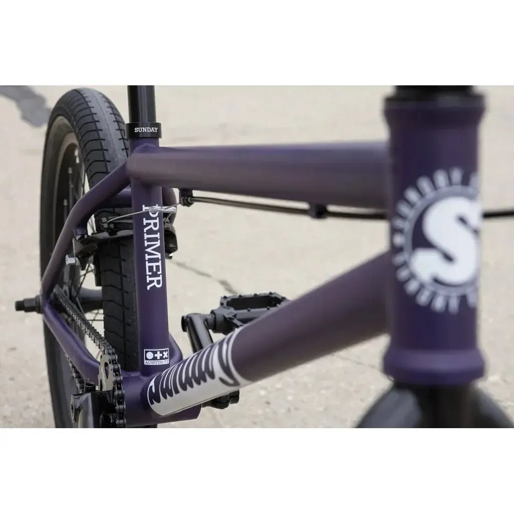 Close-up of a purple Sunday Primer 20 Inch Bike frame, featuring the "PRIMER" label and rear wheel, with visible "Sunday" branding.