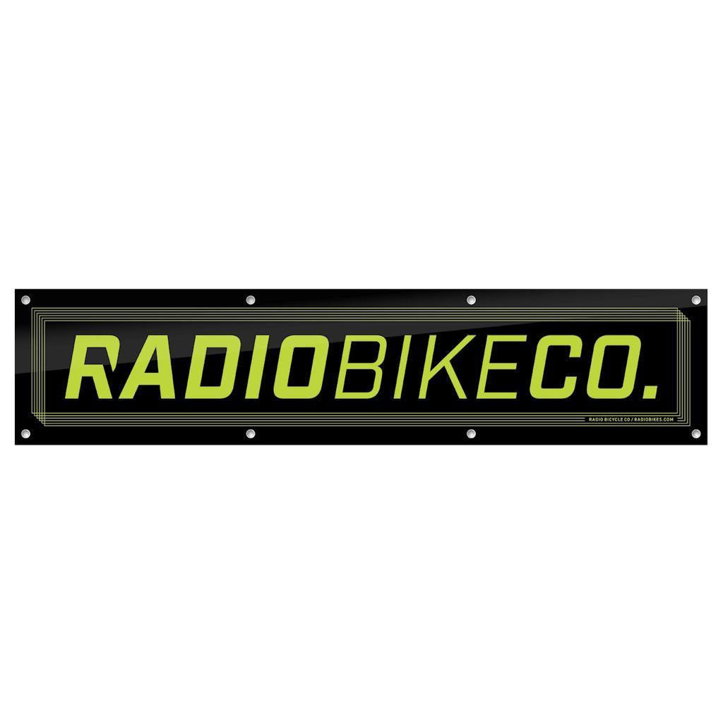 The Radio Contest Banner is a rectangular black PVC sign with yellow-green "RADIOBIKECO." text and edge mounting holes—ideal for outdoor use or displaying your favorite BMX brand.