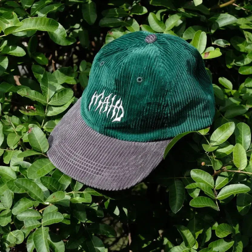 The Mahd Metal Corduroy Hat, featuring green and gray corduroy with white embroidered text, rests on leafy shrubs—its crown distinct against the foliage.