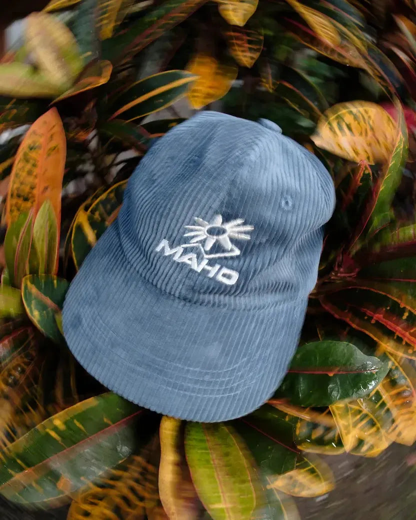 The Mahd Botanical Corduroy Hat, a blue cap featuring a white sun and “MAHD” logo, adds standout style as it sits atop colorful, variegated leaves.